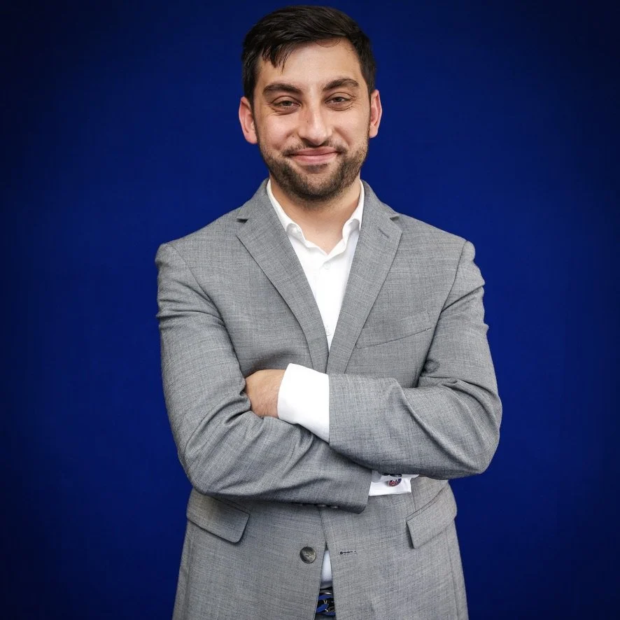 A man in a gray suit with crossed arms, standing against a dark blue background.