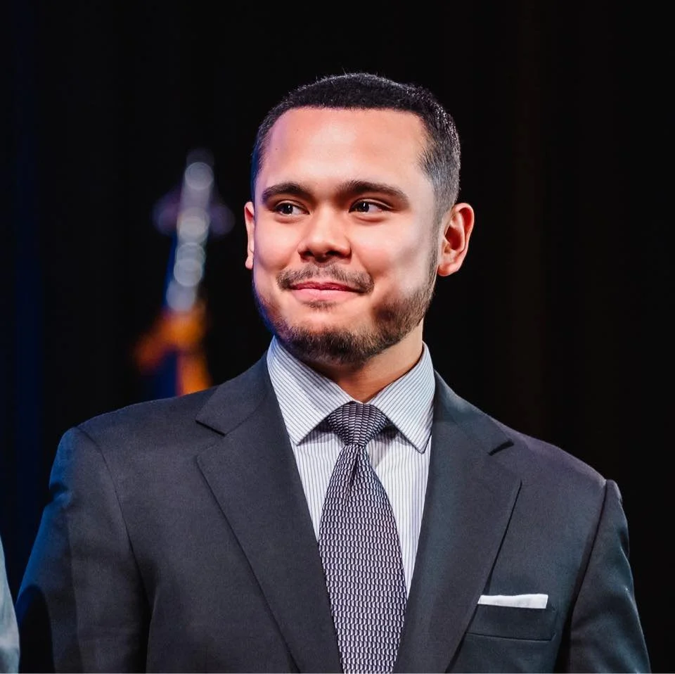 A young man in a dark suit with a tie, smiling softly, standing in front of a dark background with a blurred flag.
