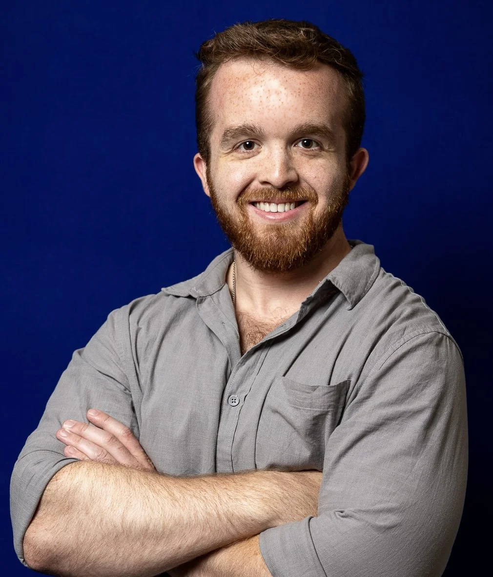 A smiling man with red-brown hair and a beard, wearing a gray shirt, poses with arms crossed against a blue background.