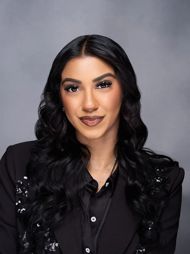 An Indian woman with long, black, wavy hair, wearing a black blazer with sequin accents, looking at the camera, set against a gray background.