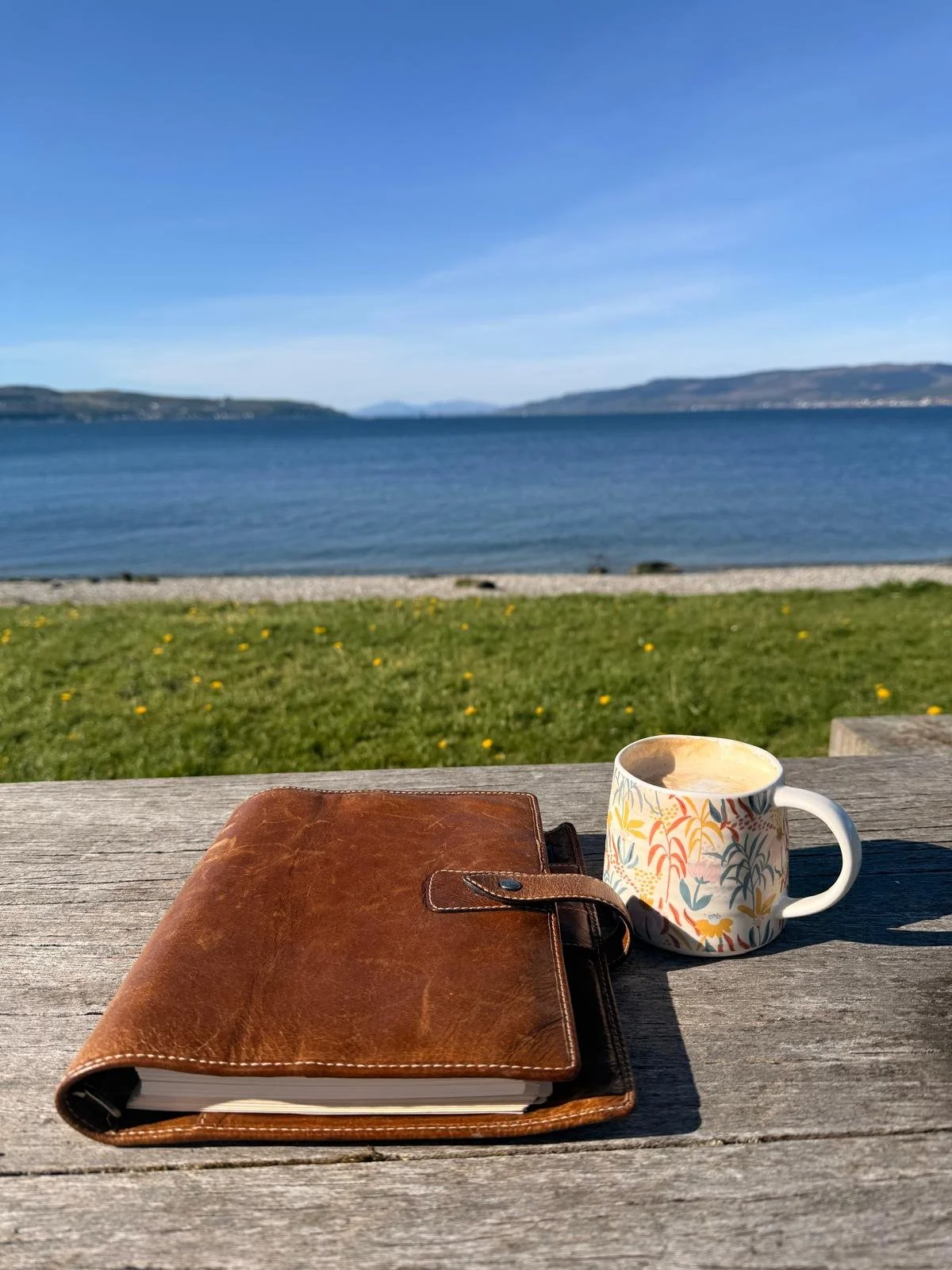 A leather-bound notebook, a ceramic mug with a floral pattern, and a small wooden table with a scenic lake view, mountains, blue sky, and grassy area with yellow flowers.