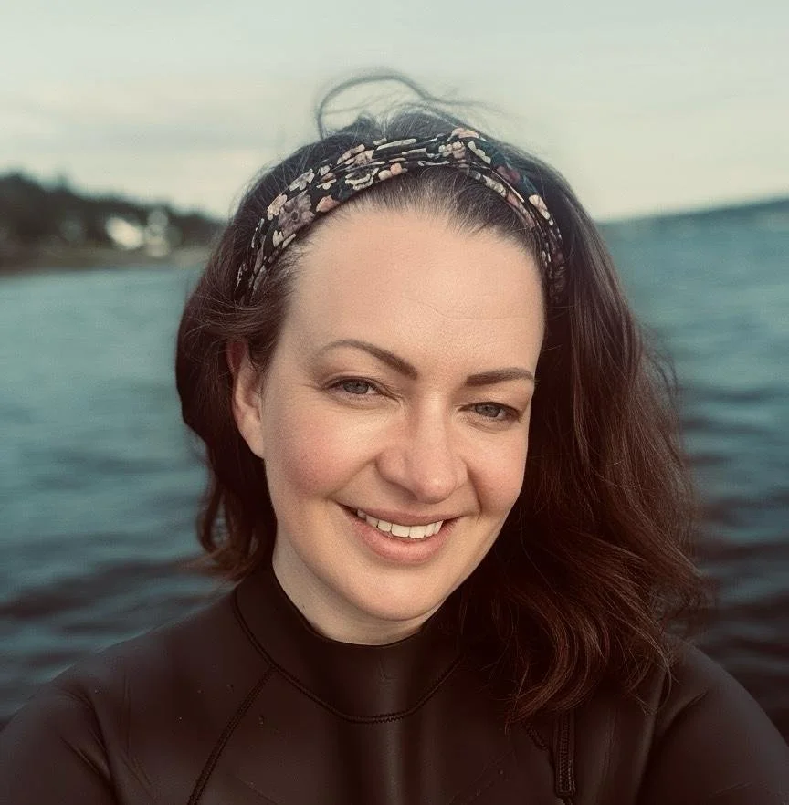 A young woman with dark hair wearing a headband and a black top, smiling while near a body of water with land and houses in the background.