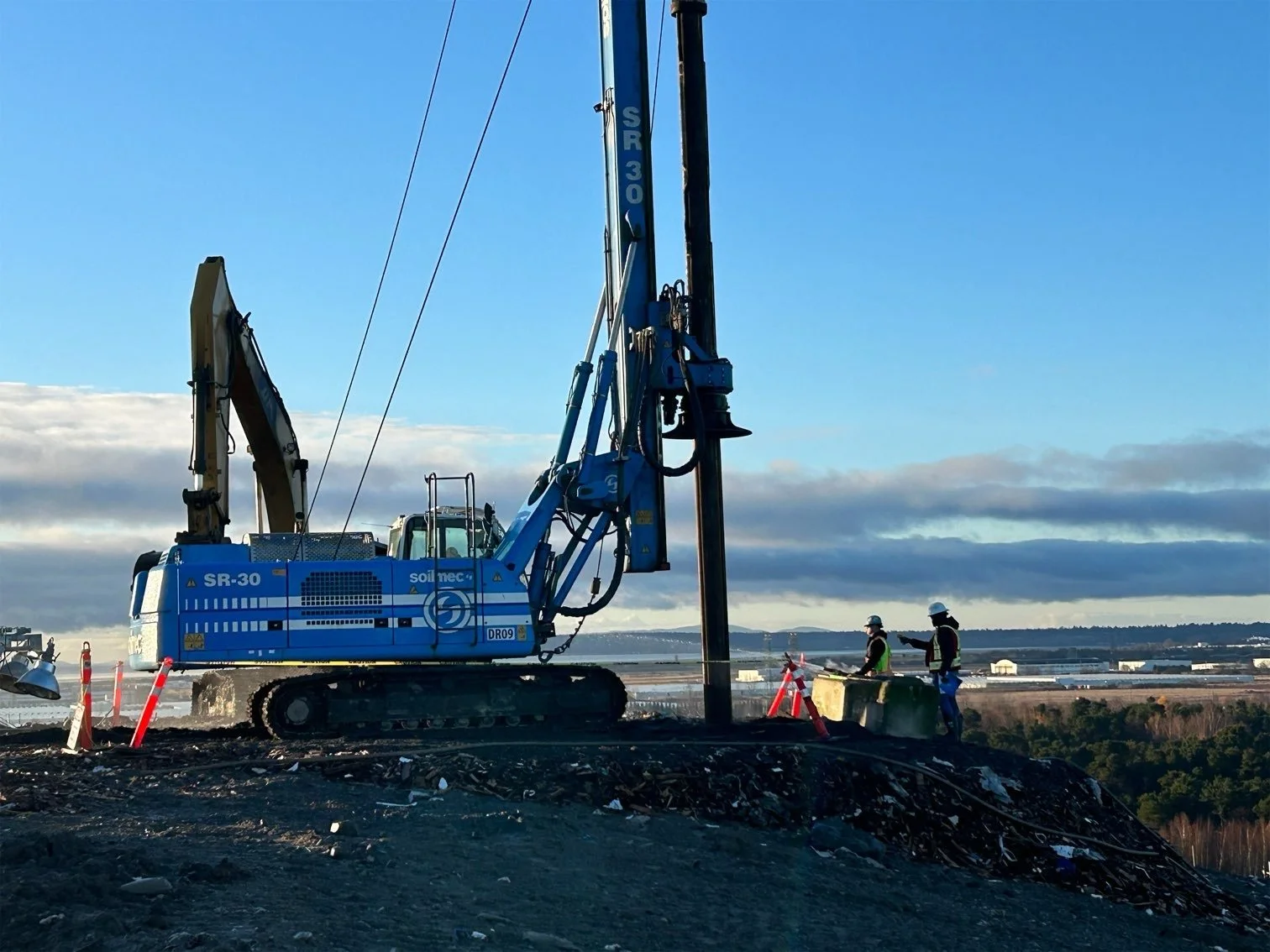 Construction workers in safety gear operating heavy machinery, including an SR-30 drilling rig, on a construction site with a scenic background of sky and distant landscape.