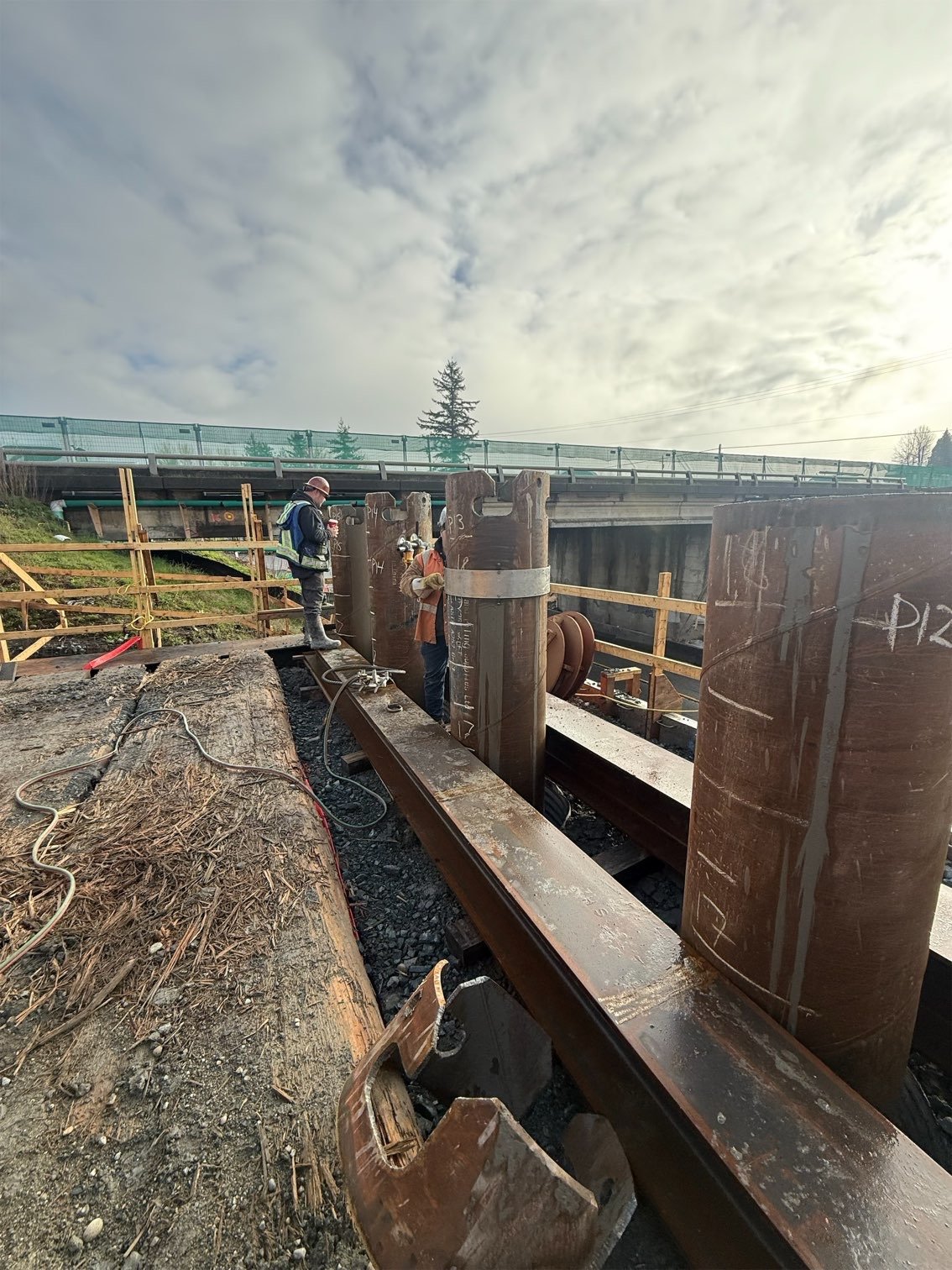 Two construction workers are working on installing a segment of railroad track under a bridge, with safety barriers around the area and a cloudy sky above.