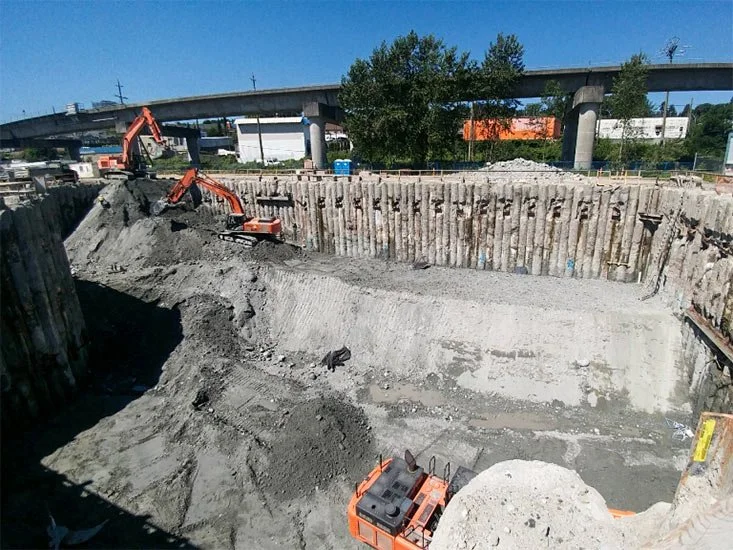 Construction site with excavators digging a large hole, surrounded by wooden support beams, with overpass and trees in the background.