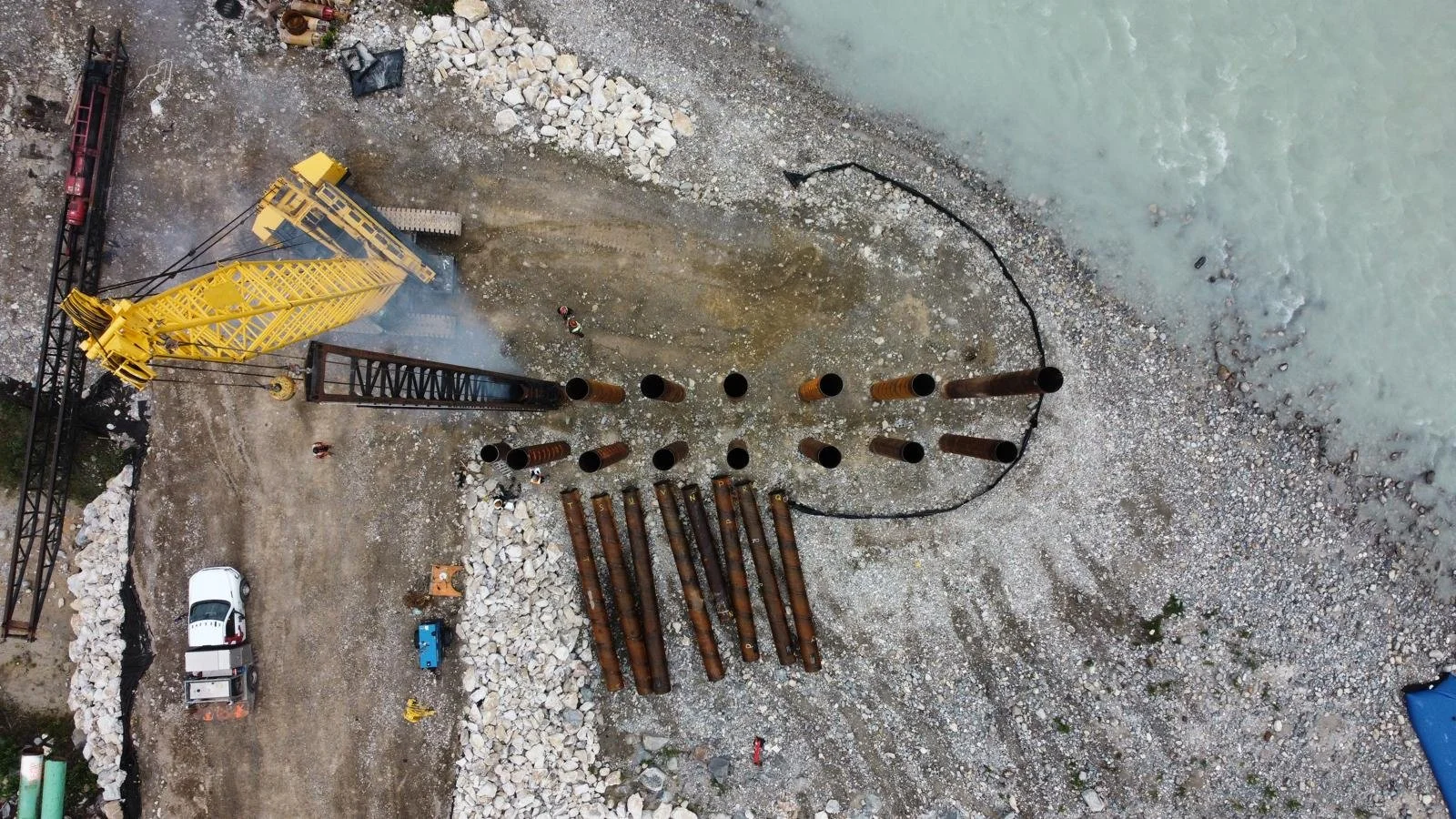 An aerial view of a construction site near a body of water, showing large pipes, construction workers, a yellow crane, and construction vehicles.