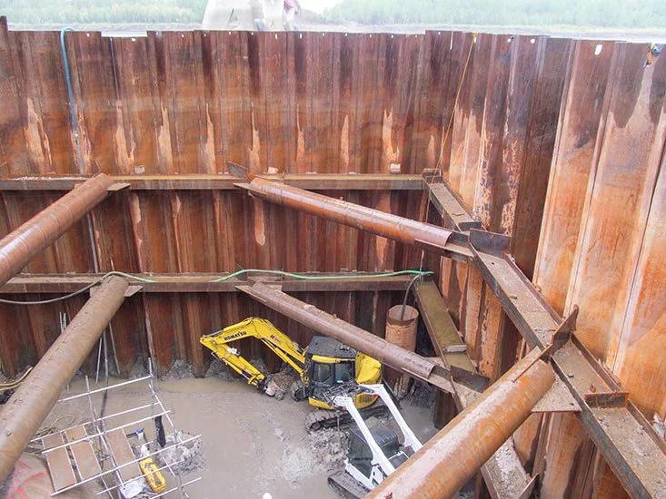 Construction site with a deep excavation supported by large steel beams and pipes, with a small excavator at the bottom.