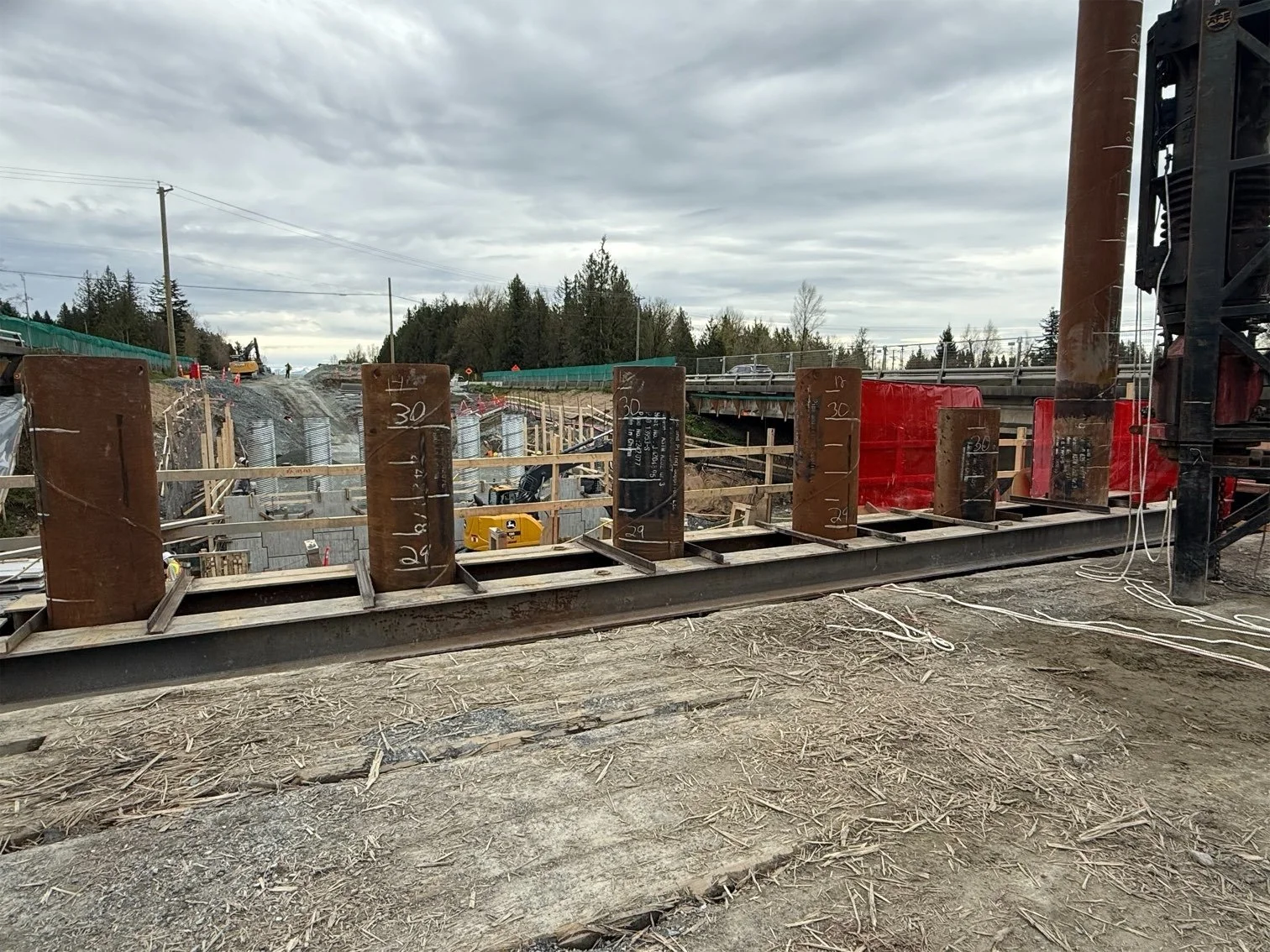 Construction site with large steel beams and wooden formwork for concrete pouring, overcast sky, trees in background, construction equipment and materials visible.