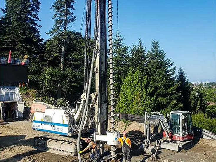 Construction site with a large pile driver and excavator, two workers in safety vests and helmets, dirt ground, surrounded by trees and a clear sky.
