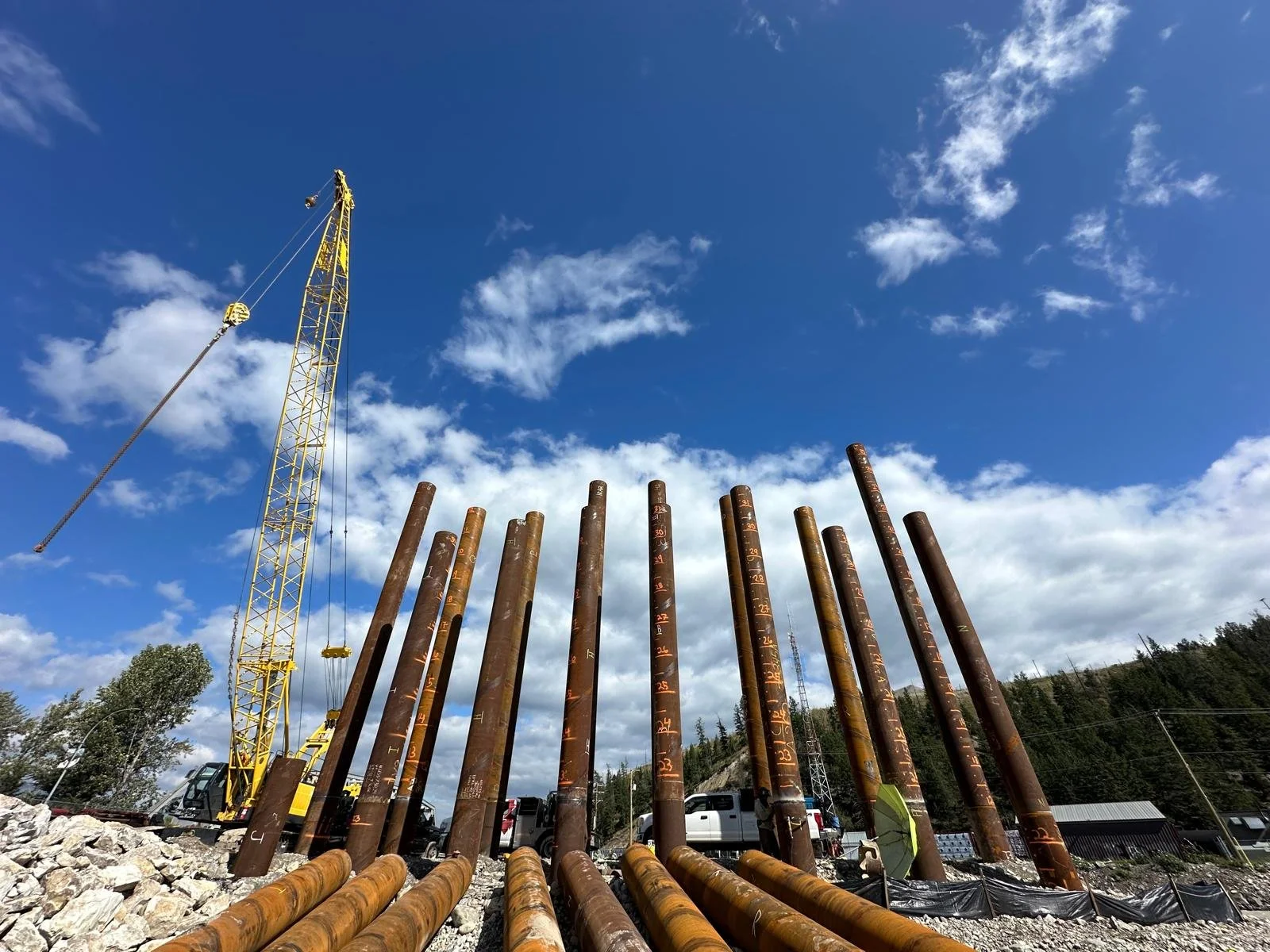 Construction site with a yellow tower crane and tall rusted metal pipes marked with numbers, set against a blue sky with clouds.