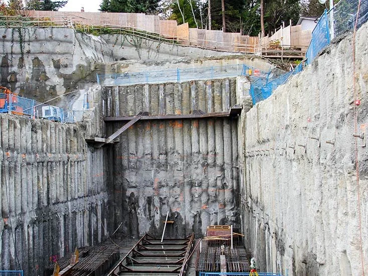 Construction site with excavated deep trench supported by steel reinforcing and retaining wall. Wooden planks and construction equipment are at the bottom.