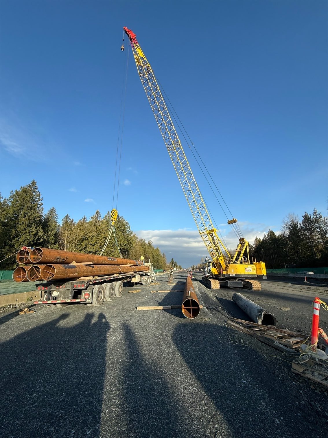 Construction site on a roadway with a large yellow crane lifting metal pipes onto a flatbed truck, trees in the background, and a clear blue sky.