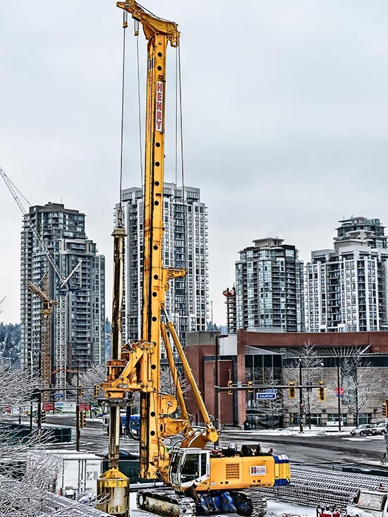 Yellow construction crane on a city street with tall modern apartment buildings in the background, during winter weather.