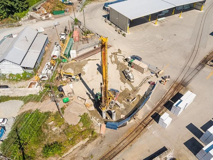 An aerial view of a construction site with a yellow drilling machine, scattered construction materials, and nearby parked vehicles and buildings.