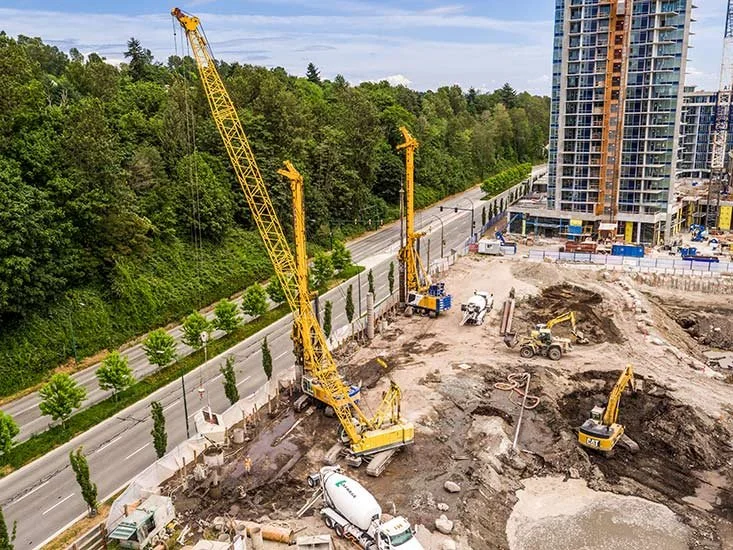 Construction site with cranes, excavators, and a cement mixer, surrounded by a multi-story building and trees.