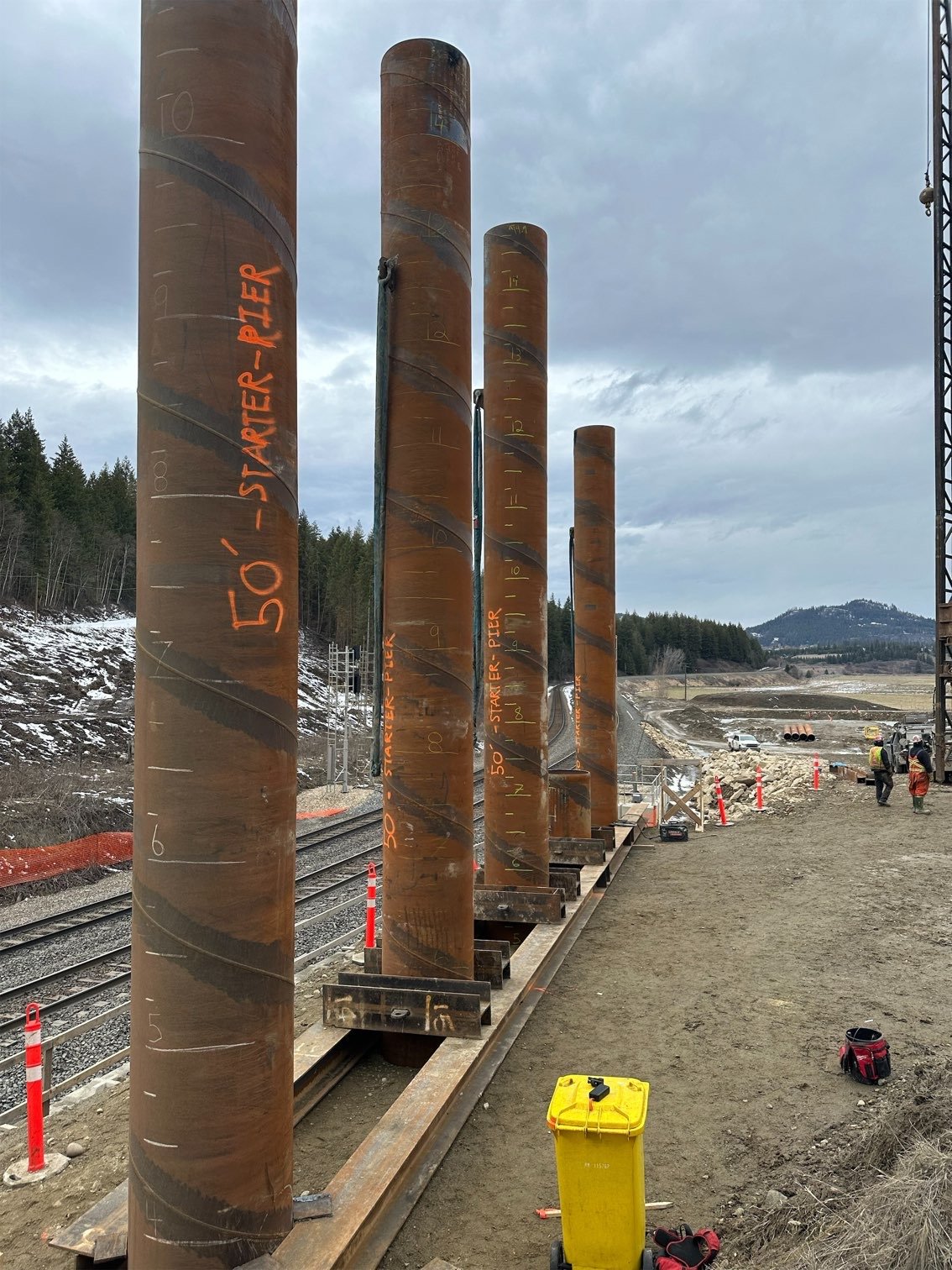 Four large rust-colored steel pipes standing vertically on a construction site next to railroad tracks with hills and cloudy sky in the background.