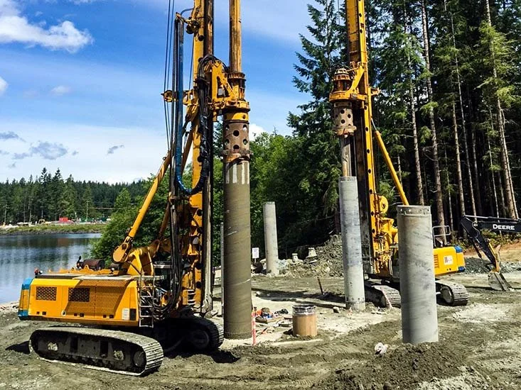 Two large yellow pile drivers on tracks positioned vertically for foundation work at a construction site near a body of water with tall trees in the background.