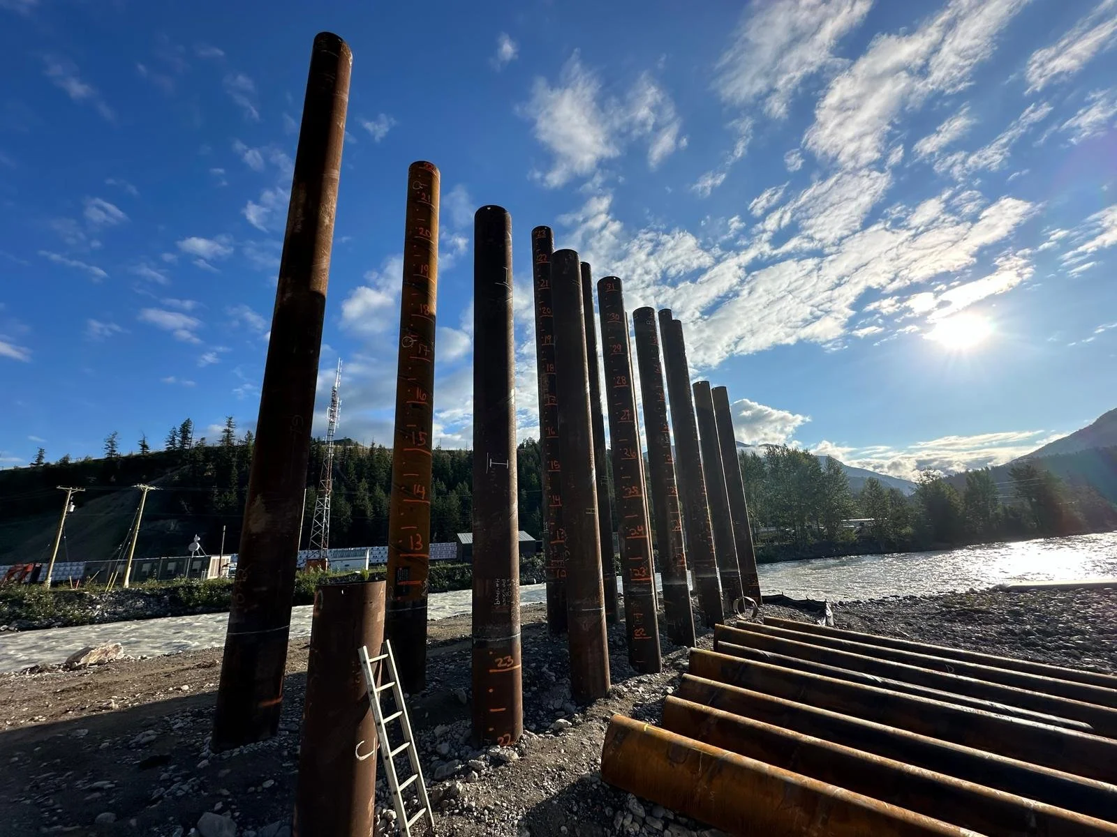 Tall steel pipes with measurement markings are lined up along a riverbank with a scenic mountain and forest landscape in the background, under a partly cloudy blue sky and bright sunlight.