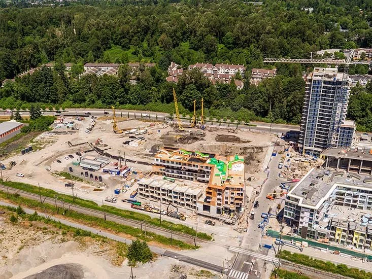 Aerial view of a construction site with cranes, partially built structures, and ongoing building work, surrounded by trees and nearby buildings.