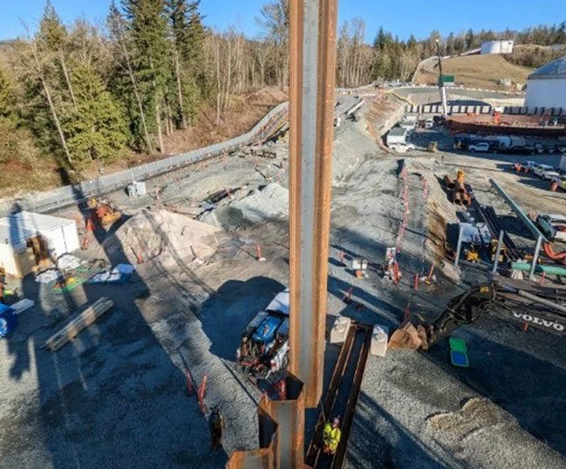 View of a construction site with heavy machinery, gravel, and poles during daytime. Some workers are visible.