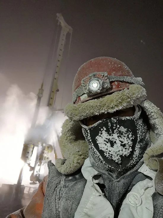 A person wearing a hard hat with a headlamp, safety goggles, a dust mask, and a winter coat, covered in frost and snow, working in a snowy, industrial environment.