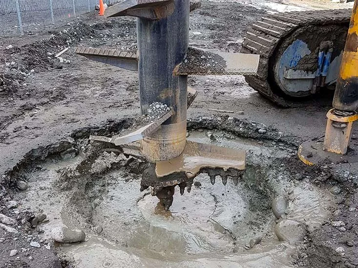 Close-up of a construction site showing an auger drill bit in the ground surrounded by wet concrete and dirt, with part of an excavator visible.