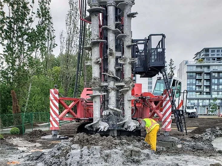 Worker in yellow safety gear inspecting large construction drill at a construction site with urban buildings and trees in the background.
