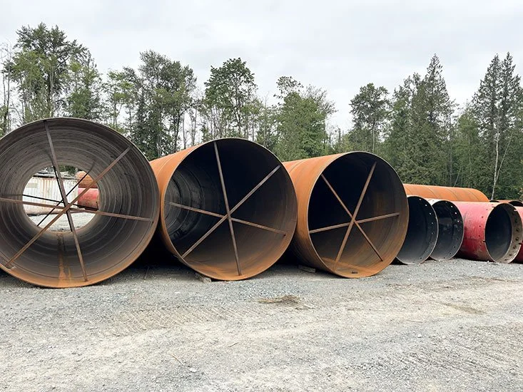 Large industrial metal pipes stacked outside on gravel, with trees and cloudy sky in the background.
