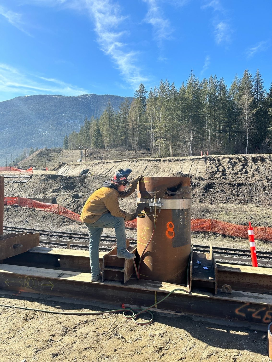 A worker at a construction site near railroad tracks, wearing a helmet and safety gear, is working on a large rusted metal pipe amidst dirt and construction materials with mountains and a clear blue sky in the background.
