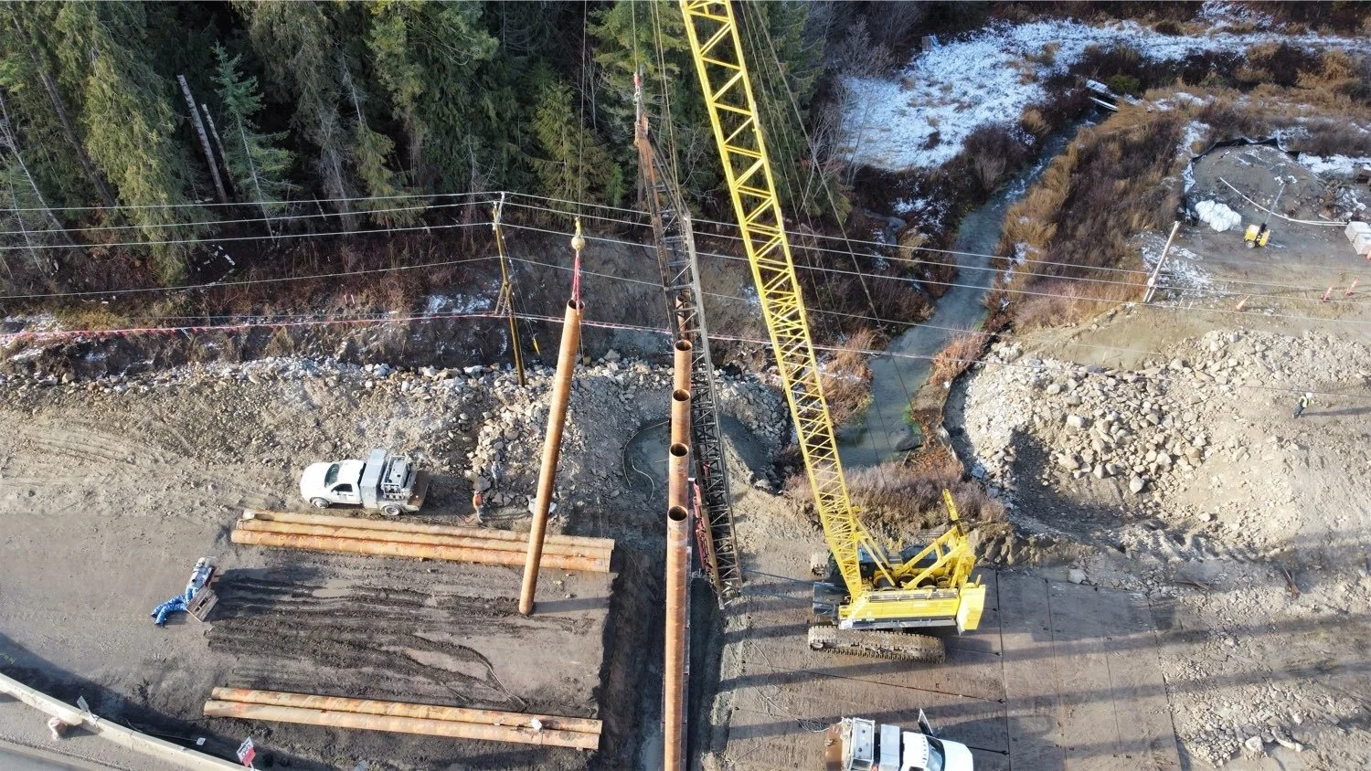 Aerial view of a construction site with a large yellow crane lifting a metal pole, dirt road, and construction workers, near a forested area and a small creek.