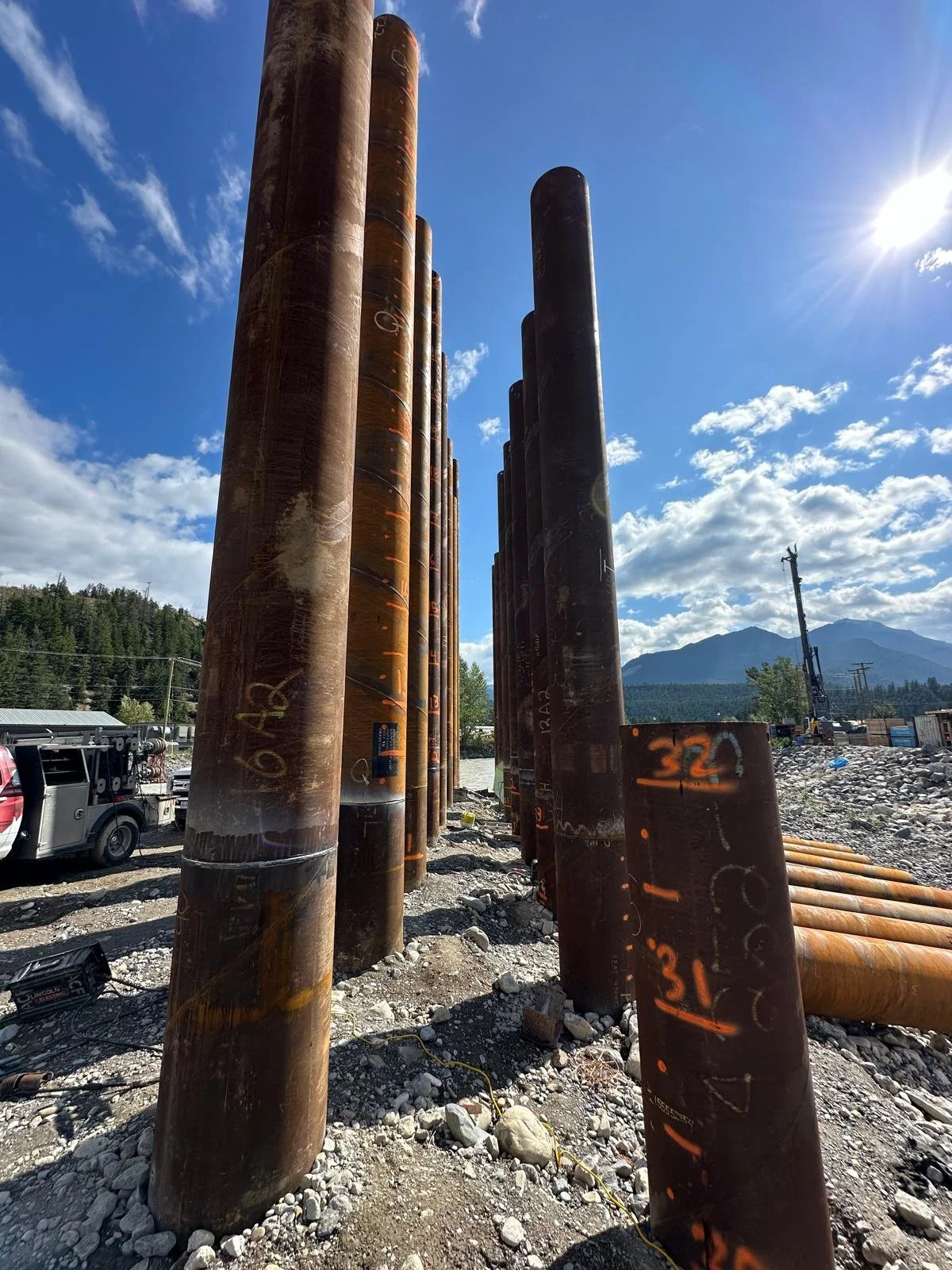 Large rusted metal pipes at a construction site with mountains in the background under a partly cloudy sky and bright sunlight.
