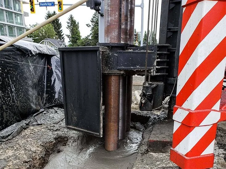 Close-up of a construction site with large machinery and equipment, including a drill or pile driver, in front of a sidewalk and street intersection with traffic lights and a street sign reading 'Hazelbridge'.