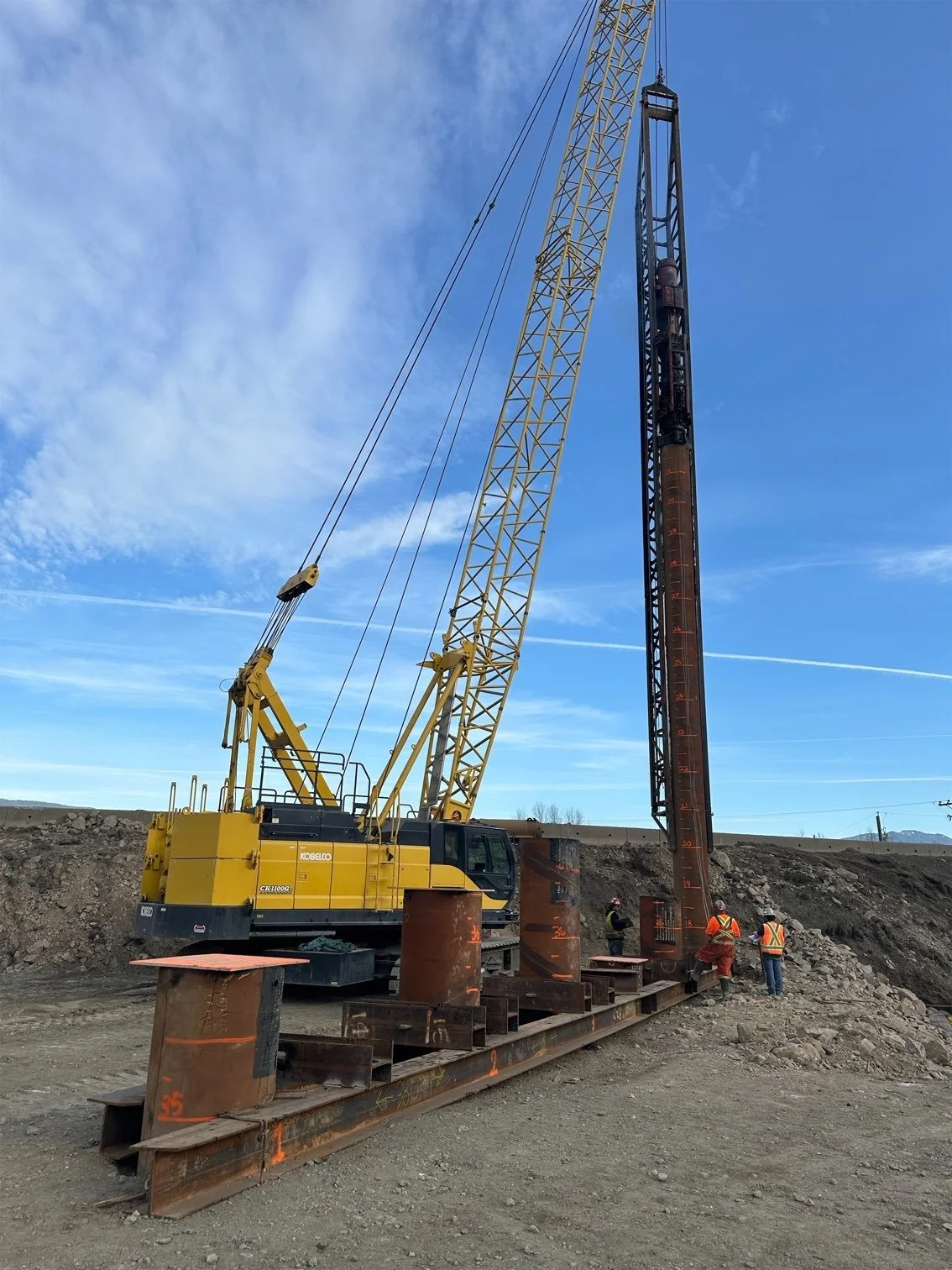 Construction site with a large yellow crane lifting a steel beam, three workers in safety vests and helmets standing nearby on a dirt ground.