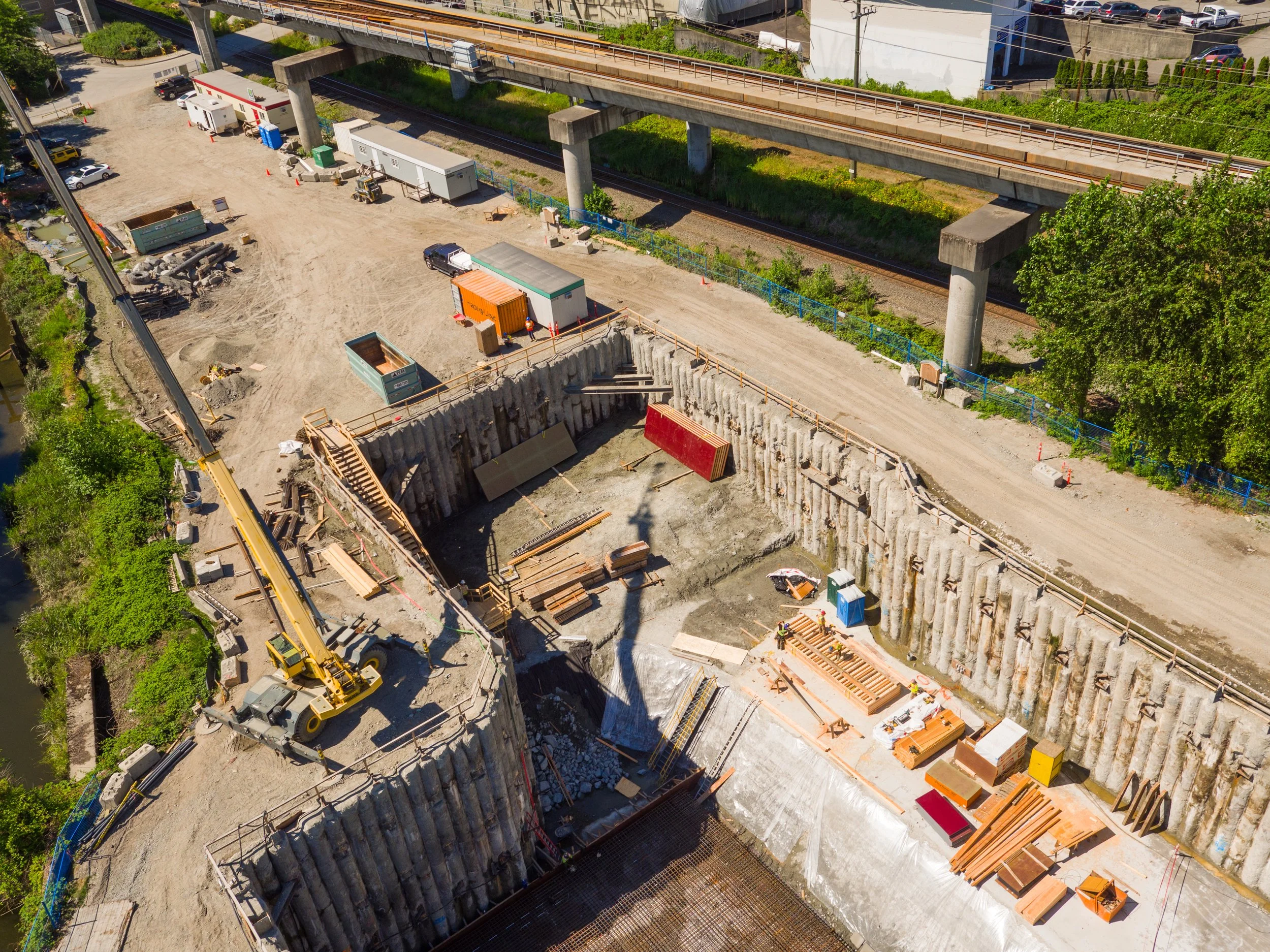 An aerial view of a construction site beneath a highway bridge, with heavy machinery, construction materials, and workers.