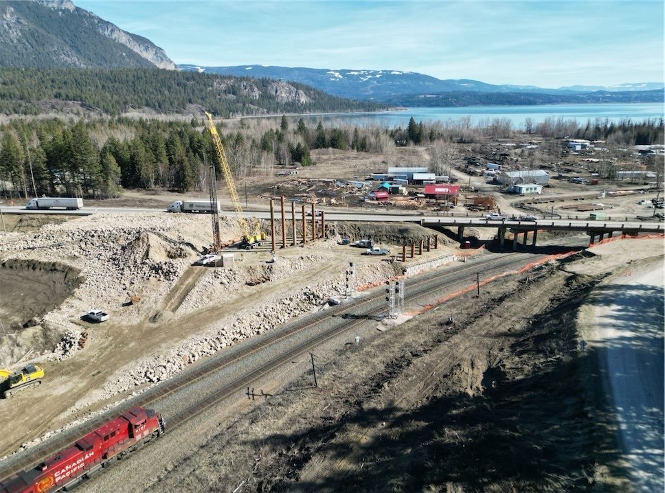 Construction site next to railroad tracks with a crane, several train cars, and a bridge, set in a rural area with trees, mountains, and a body of water in the background.