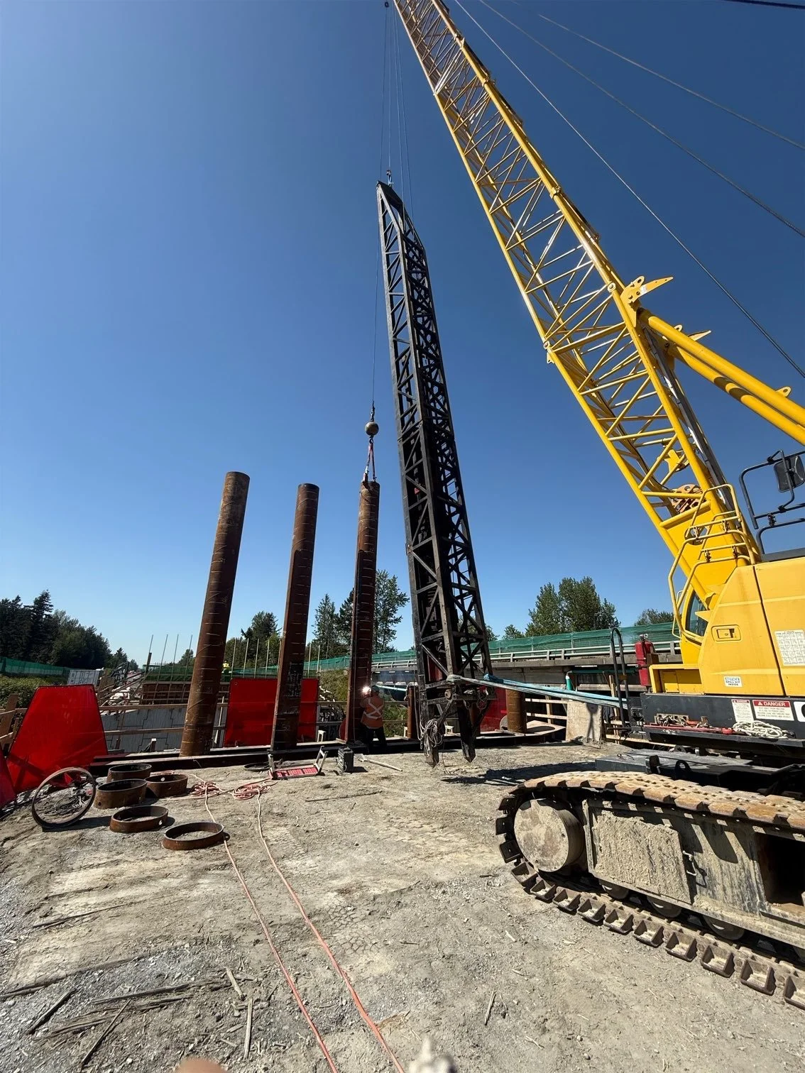 Construction site with a yellow crane and black steel structure being lifted, several rusty steel pipes, construction workers, and safety barriers under a clear blue sky.