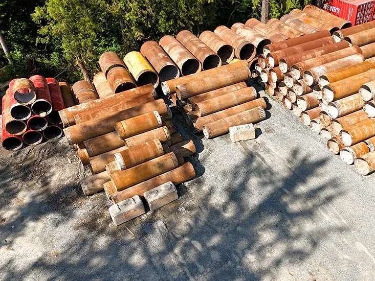 Pile of rusted large metal pipes stacked outdoors on gravel ground with some shadow from nearby trees.