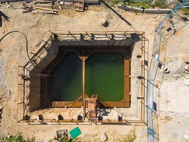 A construction site with a partially built rectangular swimming pool surrounded by dirt and construction materials, viewed from above.