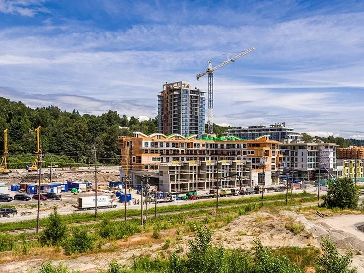 Construction site with partially built multi-story buildings, a crane, parked trucks, and green netting on some rooftops, against a background of trees and a partly cloudy sky.