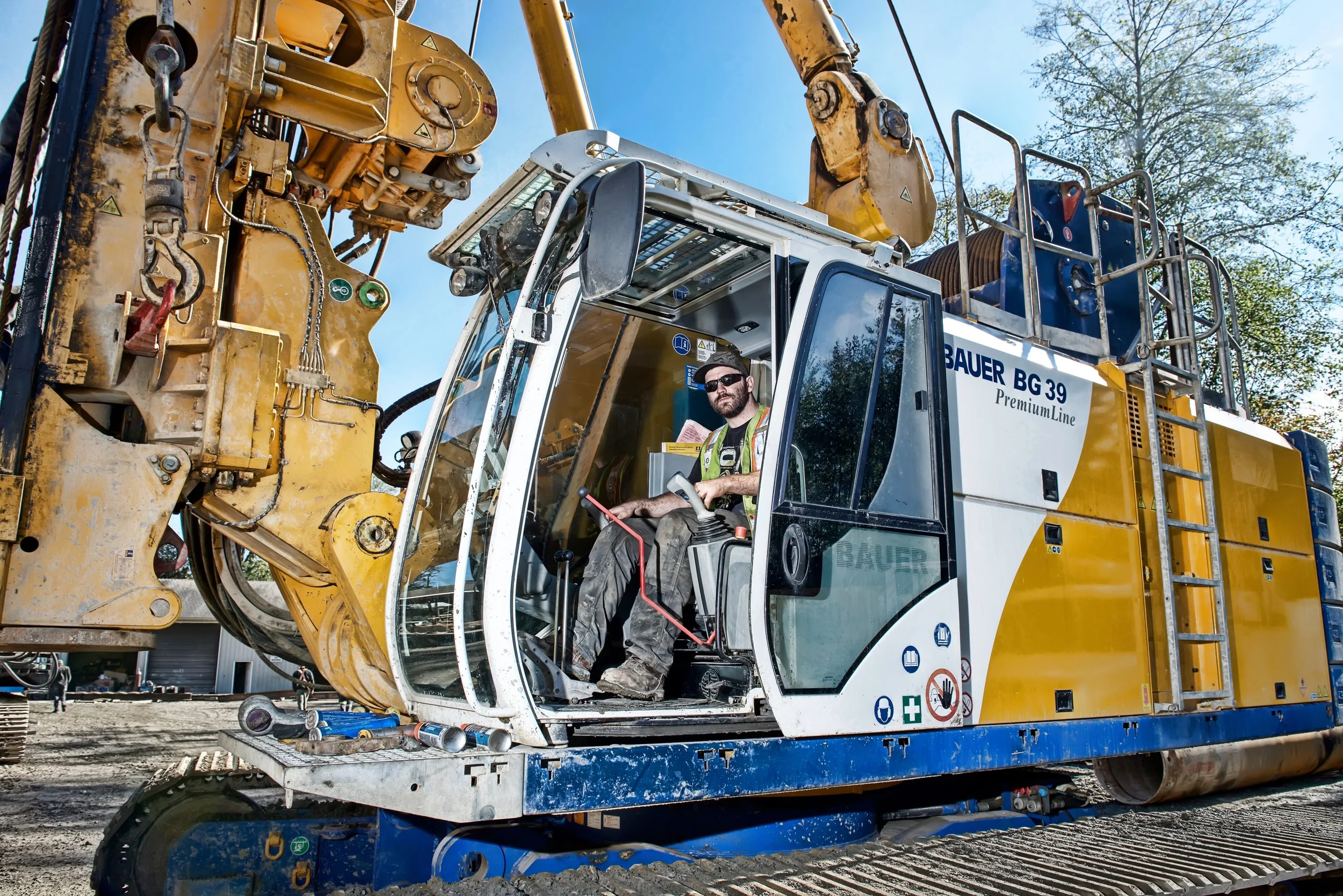 A construction worker operating a large yellow and white excavator machine outdoors on a construction site with trees and a blue sky in the background.