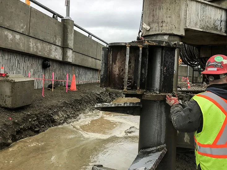 Construction worker in safety gear operating machinery at a construction site with a waterlogged area and a concrete barrier.