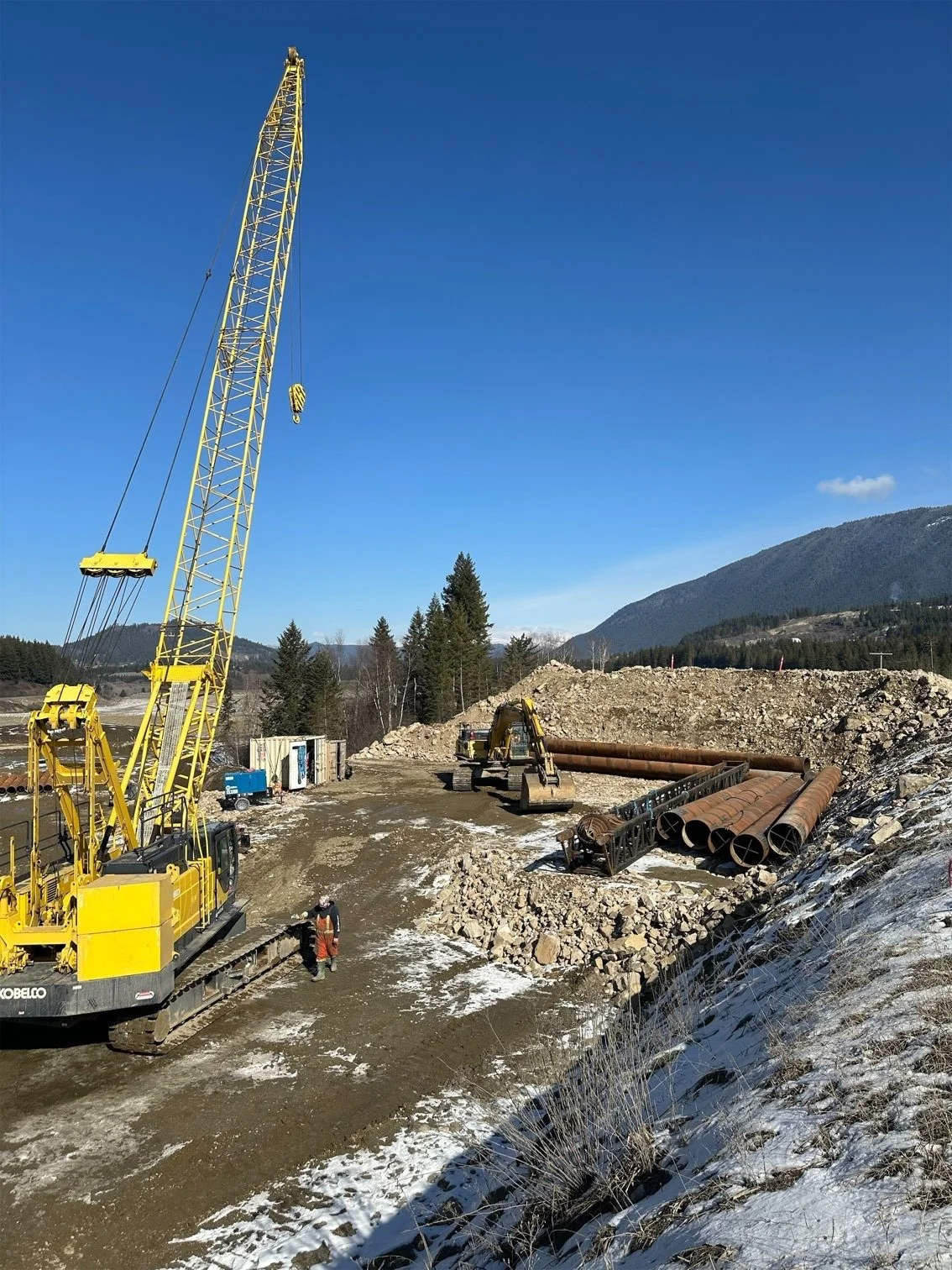 Construction site with large yellow crane, excavators, and metal pipes, set against a backdrop of mountains and trees under a clear blue sky.