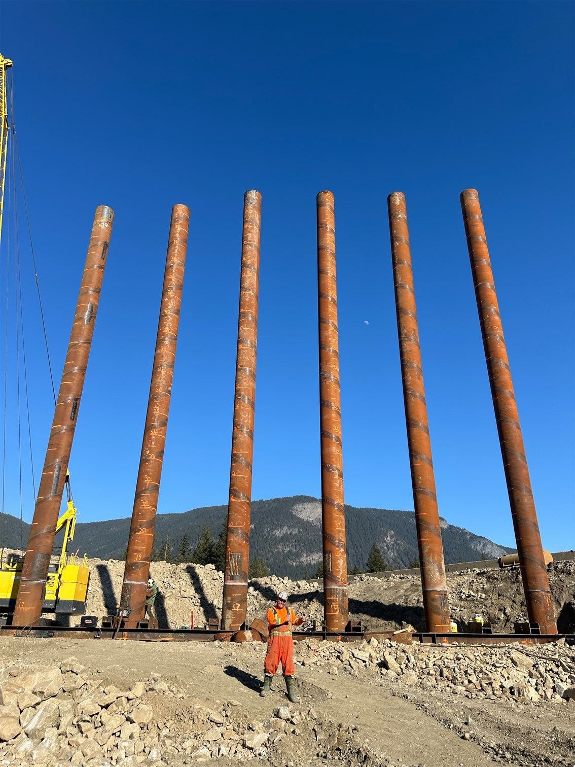 Construction site with six tall rust-colored steel columns, a construction worker in orange safety gear, mountainous background, clear blue sky with visible moon.