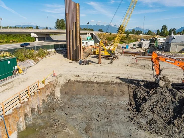Construction site with machinery, excavated earth, and an overpass in the background.