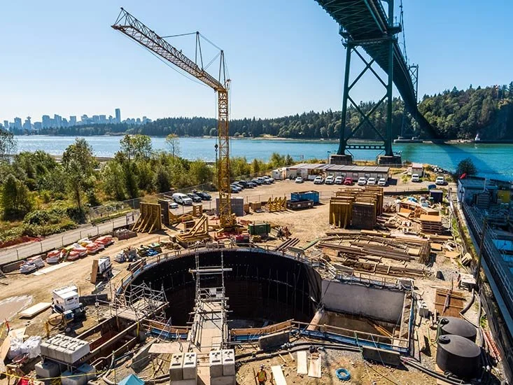 Construction site near a body of water with a large yellow crane, scaffolding, and building materials, with a bridge overhead and a city skyline in the distance.