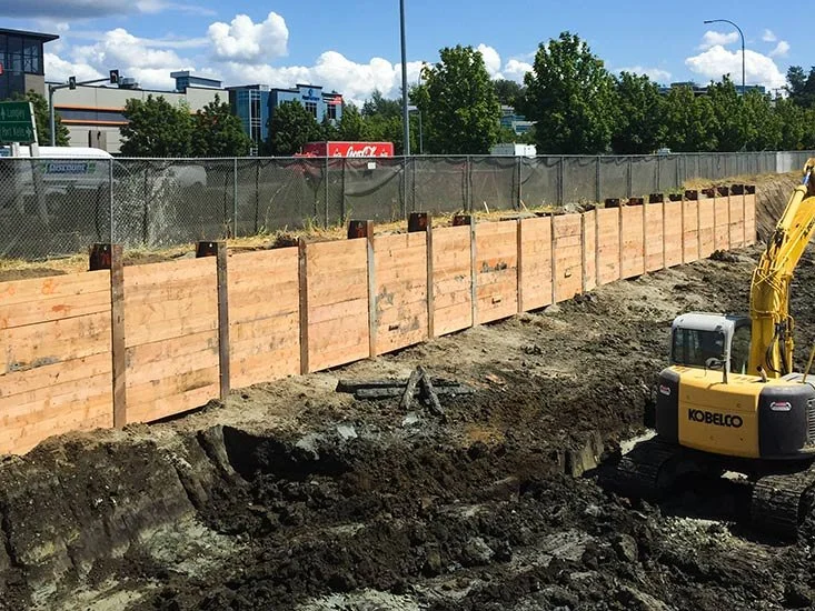 Construction site with a wooden barrier, dirt, and an excavator. Buildings, trees, and a blue sky with clouds are in the background.