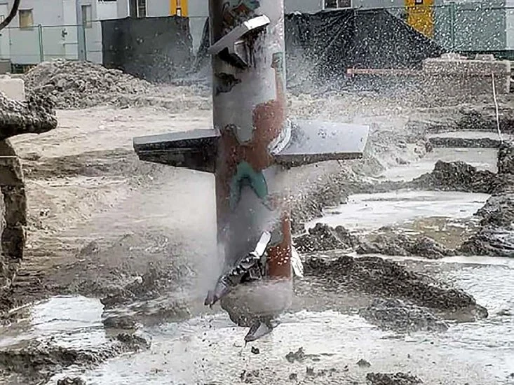 Construction site with debris and water, showing a damaged metal pole and overturned bollard in a messy environment.