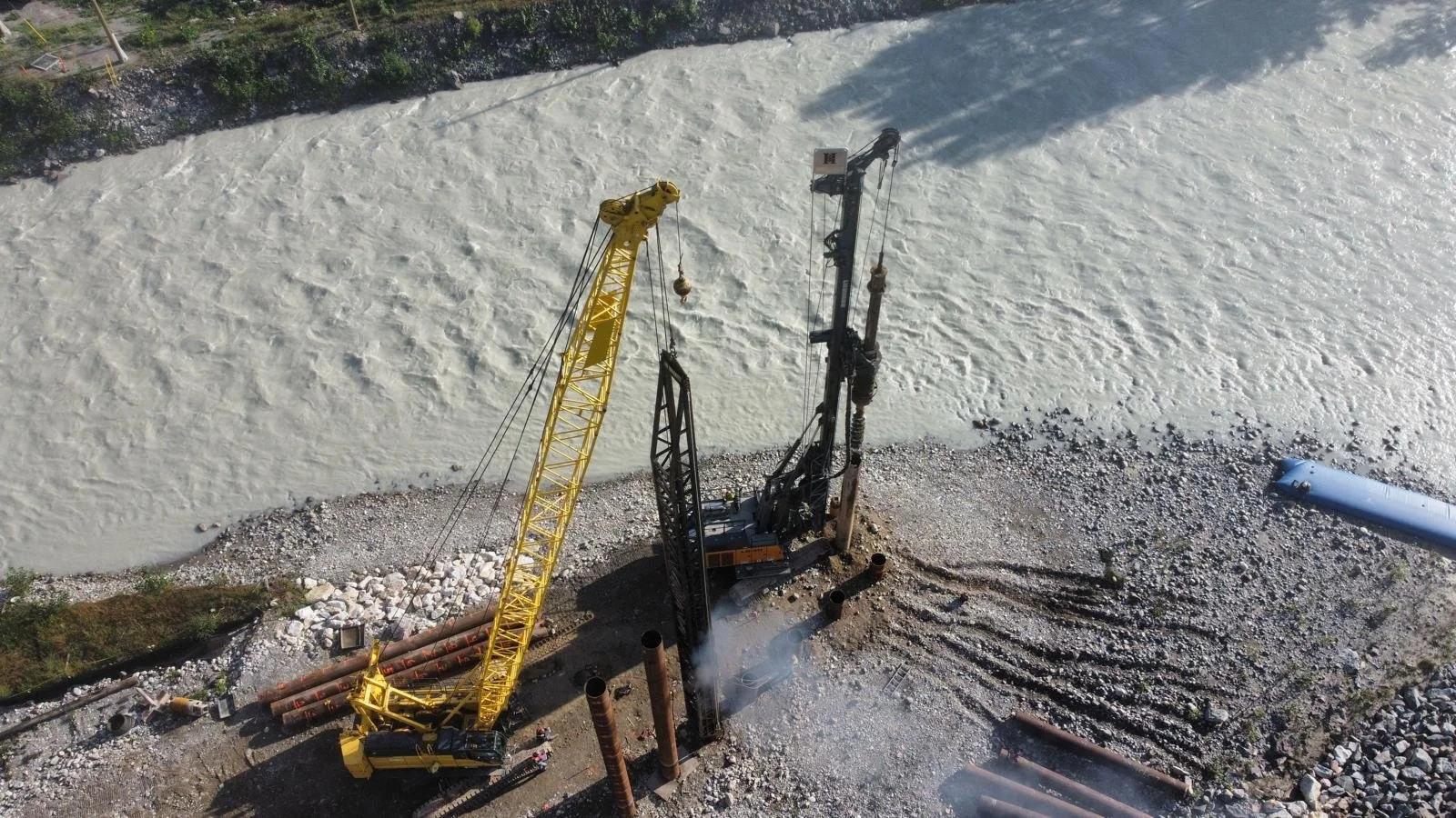 Aerial view of construction site with a yellow crane and drilling machine near a river, with pipes and rocks scattered around.