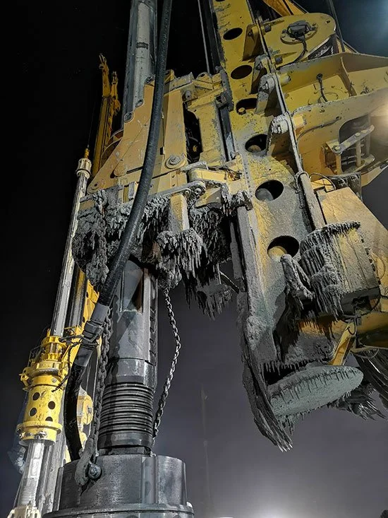 Close-up of a massive yellow construction machine with worn and torn rubber parts, exposed hydraulic components, and chains, set against a dark background.