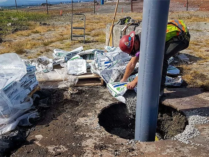 Person installing or repairing a utility pole next to a hole in the ground, surrounded by bags of supplies and construction materials, on a dry, grassy area with fencing in the background.